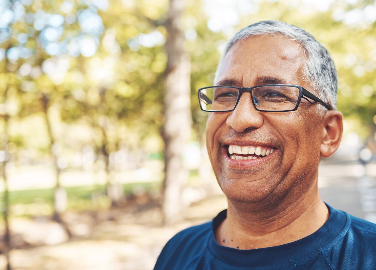 Un hombre mayor con lentes sonriendo al aire libre, bajo una luz suave.