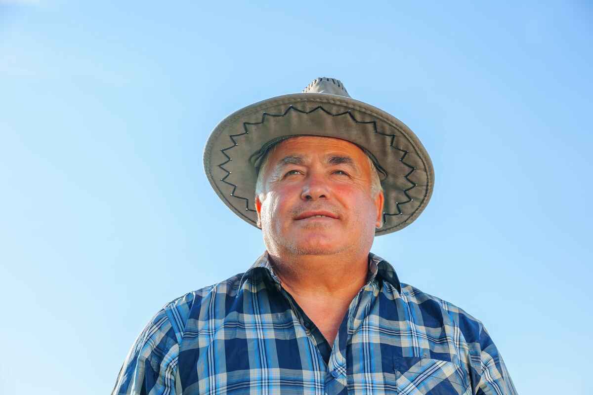 Un hombre mayor con sombrero y camisa azul a cuadros al aire libre, tranquilo y sereno.