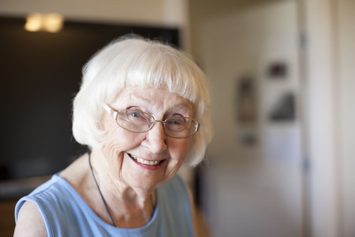 A senior woman with white hair sitting indoors, looking thoughtfully toward a window.